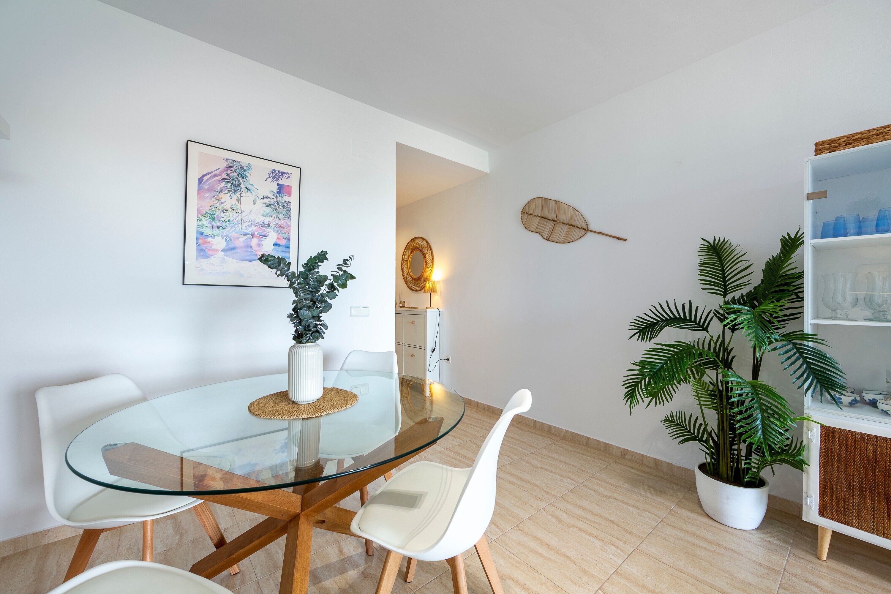 A modern dining area with a glass table, white chairs, a potted plant, and a wall-mounted shelf with decorative items.