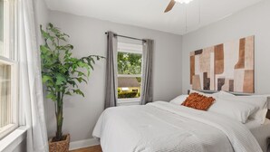 Un dormitorio con cama, ventana con cortinas, una planta en maceta y ventilador de techo.