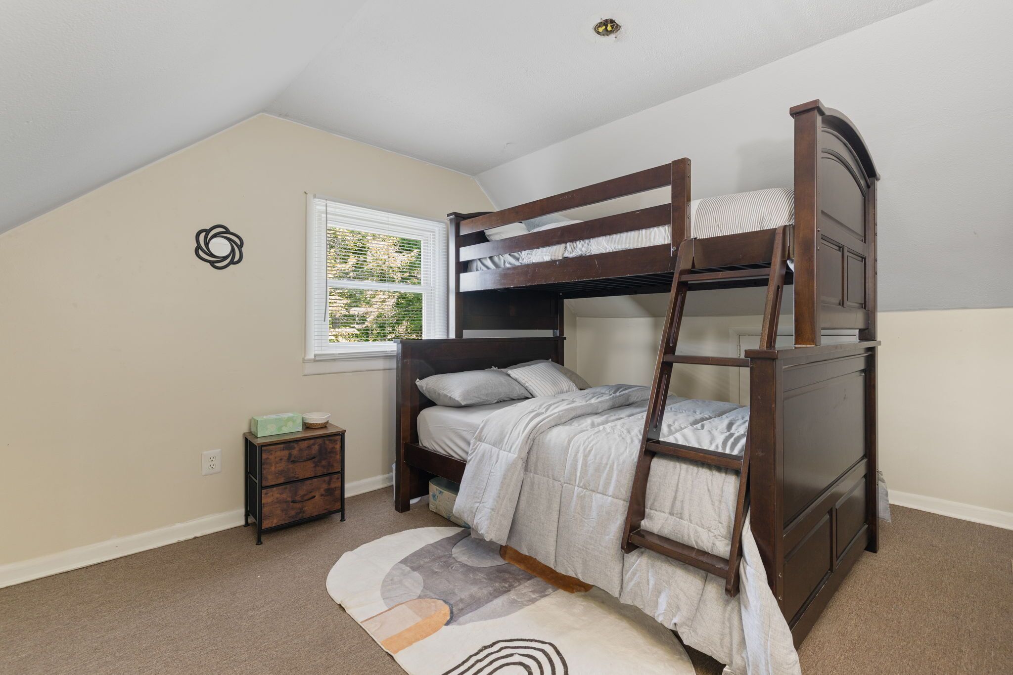 Un lit superposé dans une chambre avec une fenêtre, une petite table d’appoint en bois et un tapis à motifs.