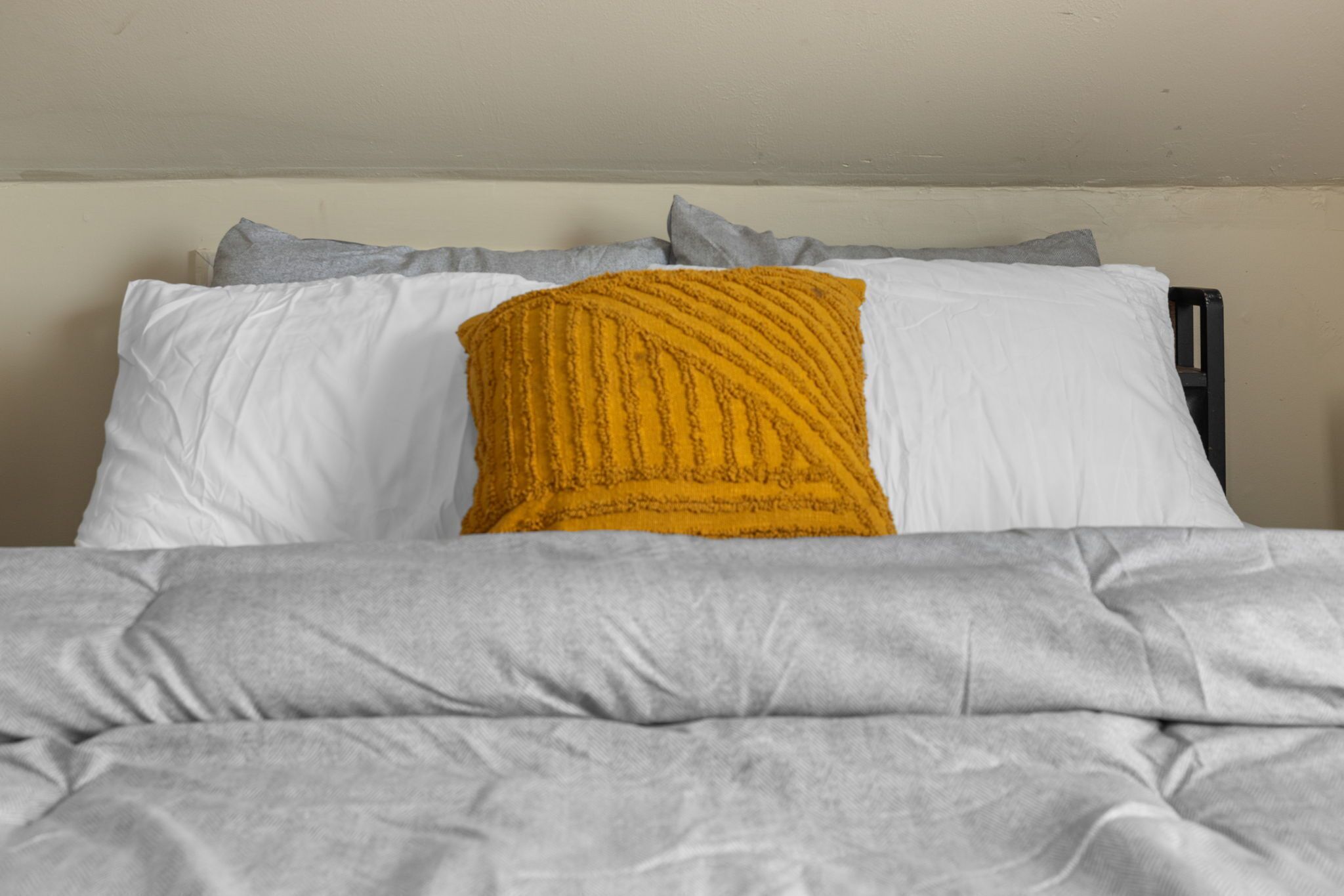 Uma cama com cobertor cinza, travesseiro branco e almofada amarela mostarda.