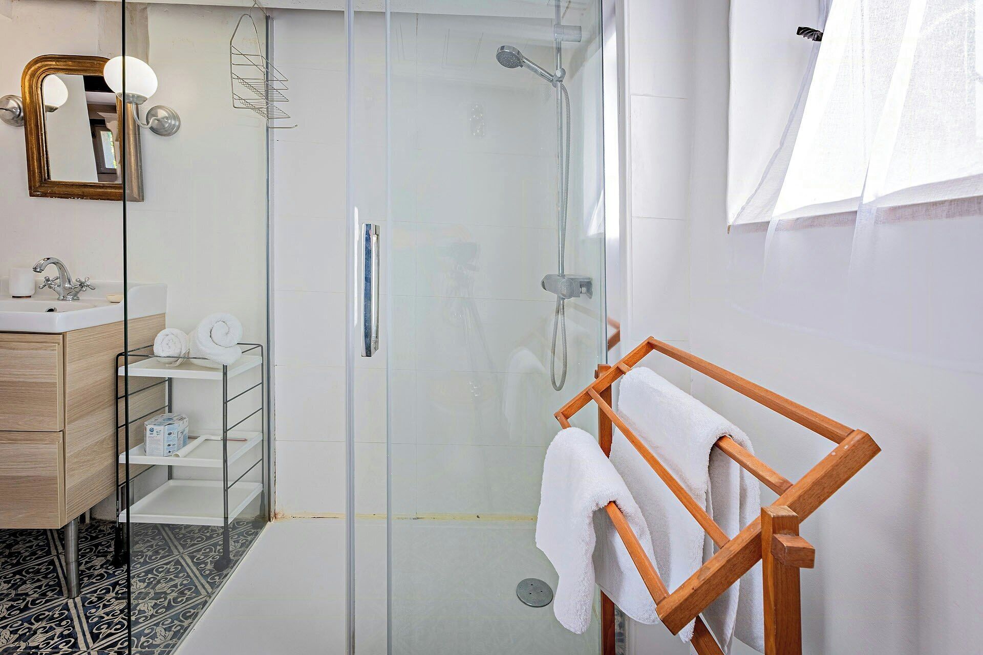 Un baño moderno con cabina de ducha de vidrio, un perchero de madera, un espejo con luces empotradas y suelo de baldosas.