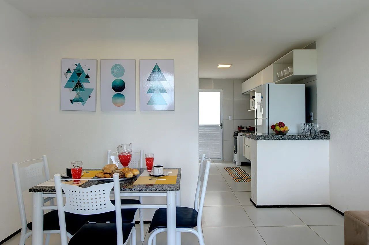 A dining area with a table set for two, a kitchen with white cabinets, and a refrigerator.