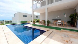 A swimming pool with a white deck, a covered outdoor seating area, and a view of a modern building. - Beach Townhouses I - Triplex by Carpediem (Aquiraz)