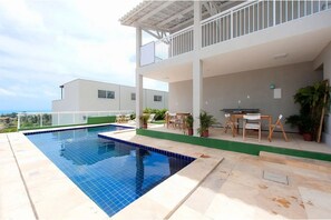 A swimming pool with a white deck, a covered outdoor seating area, and a view of a modern building. - Beach Townhouses I - Triplex by Carpediem (Aquiraz)