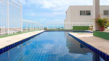 A swimming pool with clear blue tiles, surrounded by a white fence and a building with a green wall and windows.