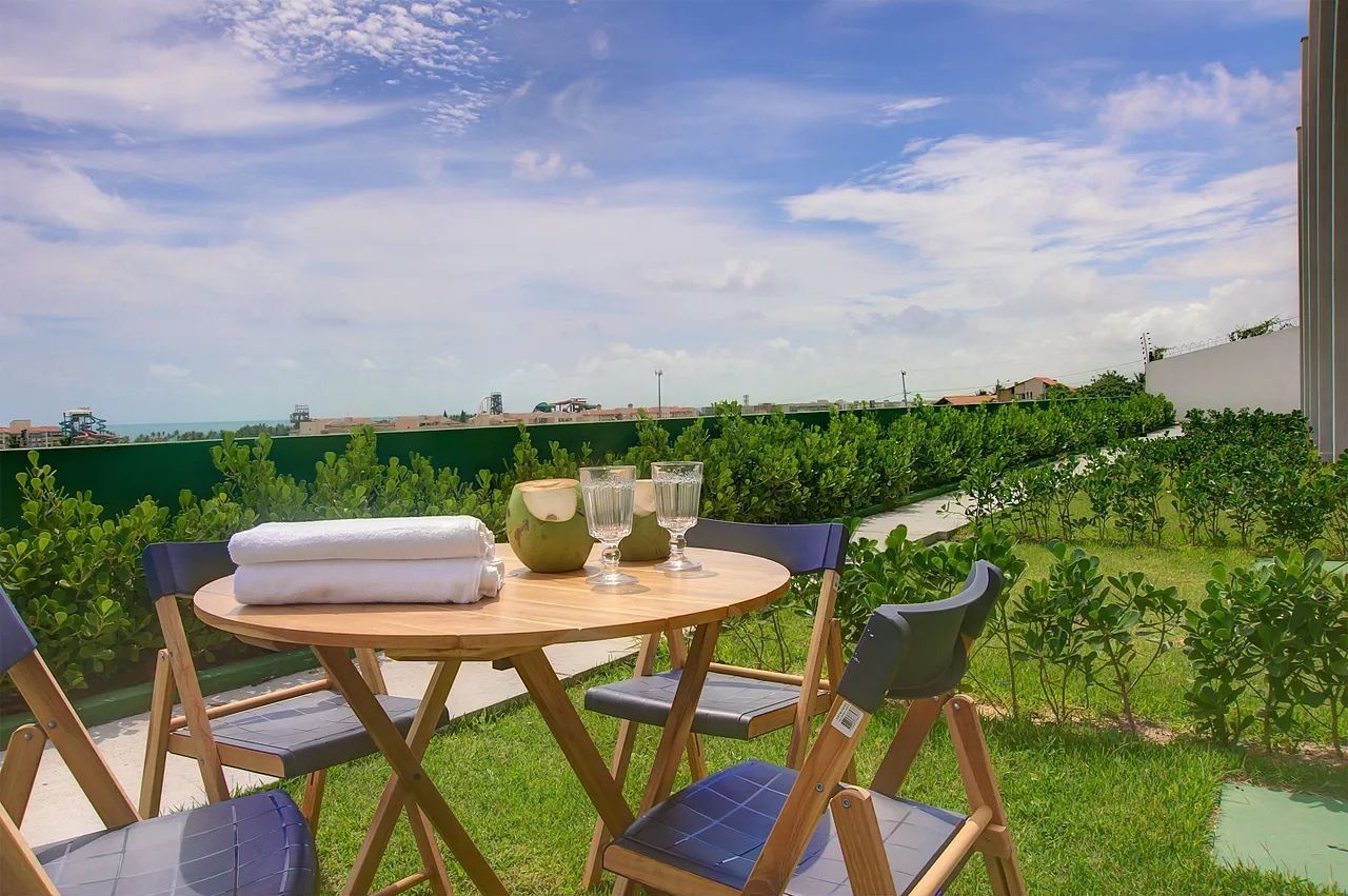 Una mesa redonda de madera con una toalla blanca y dos vasos, rodeada de sillas plegables con cojines azules, con vistas a un paisaje verde y un cielo despejado.