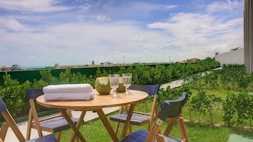 Una mesa redonda de madera con una toalla blanca y dos vasos, rodeada de sillas plegables con cojines azules, con vistas a un paisaje verde y un cielo despejado.