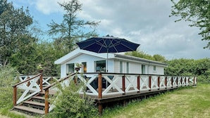 Ein weißes Hausboot mit Holzterrasse und blauem Sonnenschirm, umgeben von viel Grün.
