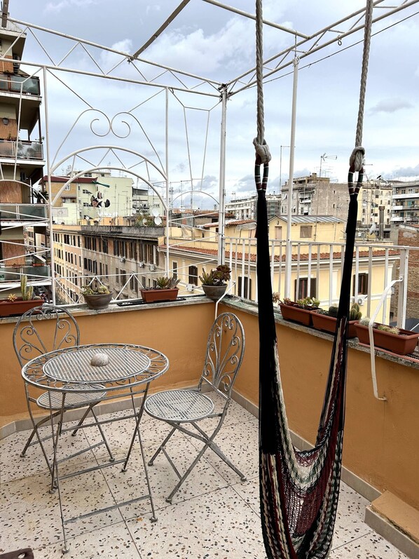 A balcony with a metal table and chairs, potted plants, and a hammock. - Small attic with romantic terrace in the heart of Torpignattara. (Roma)