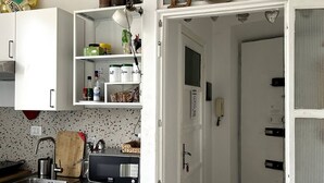 Pieni, käytännöllinen keittiö, jossa on valkoiset kaapit, mikroaaltouuni, pesuallas ja jääkaappi. Hyllyllä on purkkeja ja sen päällä koriste-esine. Ovessa näkyy sana »LOVE».