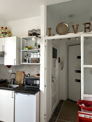 A compact kitchen with white cabinets, a microwave, a sink, and a refrigerator. There is a shelf with jars and a decorative item on top. A door with the word 'LOVE' is visible. - Small attic with romantic terrace in the heart of Torpignattara. (Roma)