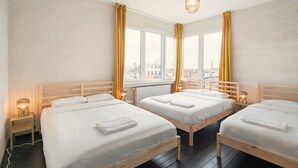 Een hotelkamer met drie houten bedden, een groot raam met gordijnen, een hanglamp en een nachtkastje met een lamp.