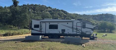 Una caravana blanca con la marca 'SHOEKUAVE' estacionada en un camino de grava, con un campo de césped y árboles al fondo.