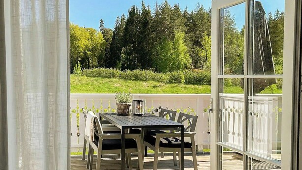 Eine Holzterrasse mit Tisch und Stühlen, eine Glastür, die nach draußen führt, und Blick ins Grüne.