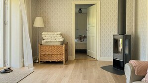 Ein gemütlicher Raum mit Kamin, einem Korbwäschesessel, einer Lampe und einer Tür zu einem weiteren Zimmer.