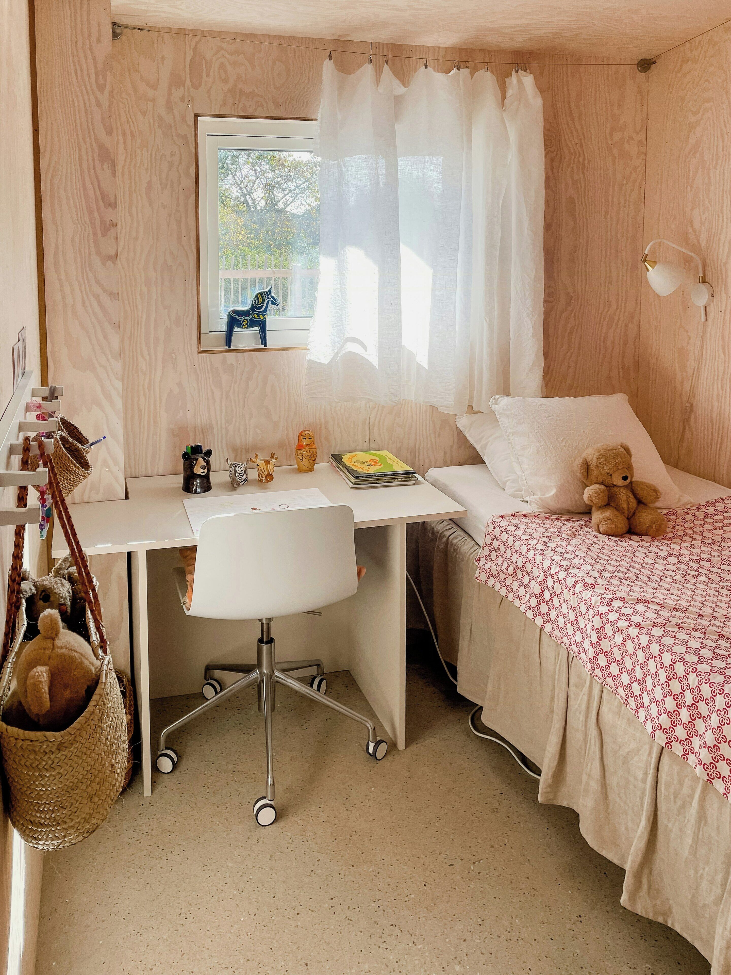 A room with a wooden desk, a bed with a white and red patterned blanket, a teddy bear on the bed, a chair, a hanging basket, and a window with sheer curtains.