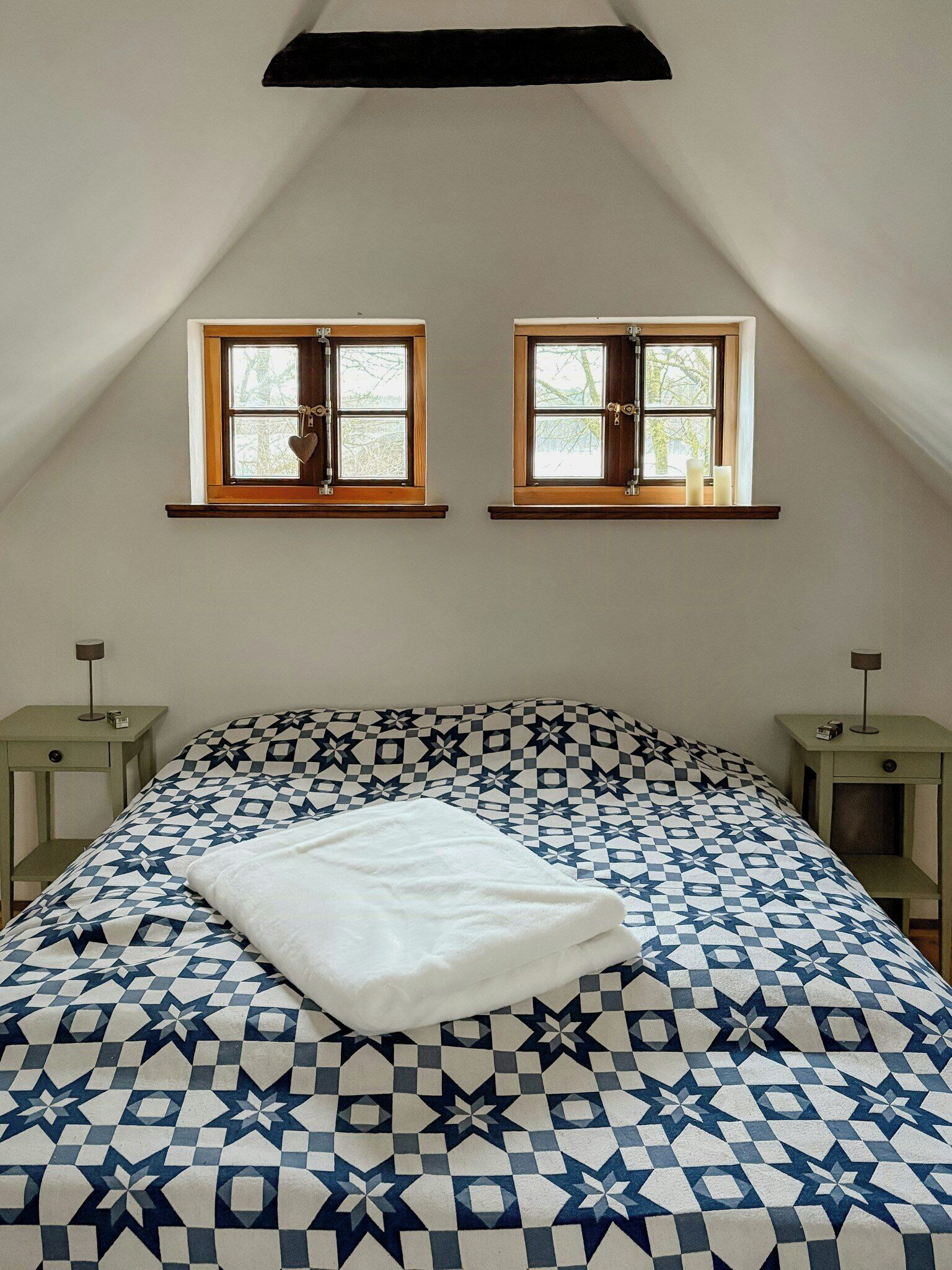 Un dormitorio con techo inclinado, dos ventanas con marcos de madera, una cama con colcha estampada y mesitas de noche con lámparas.