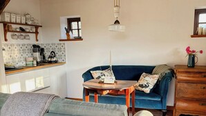 En hyggelig stue med sofa, sofabord, blå sofa, trækommode og et køkkenområde med bordplade og hængende lampe.