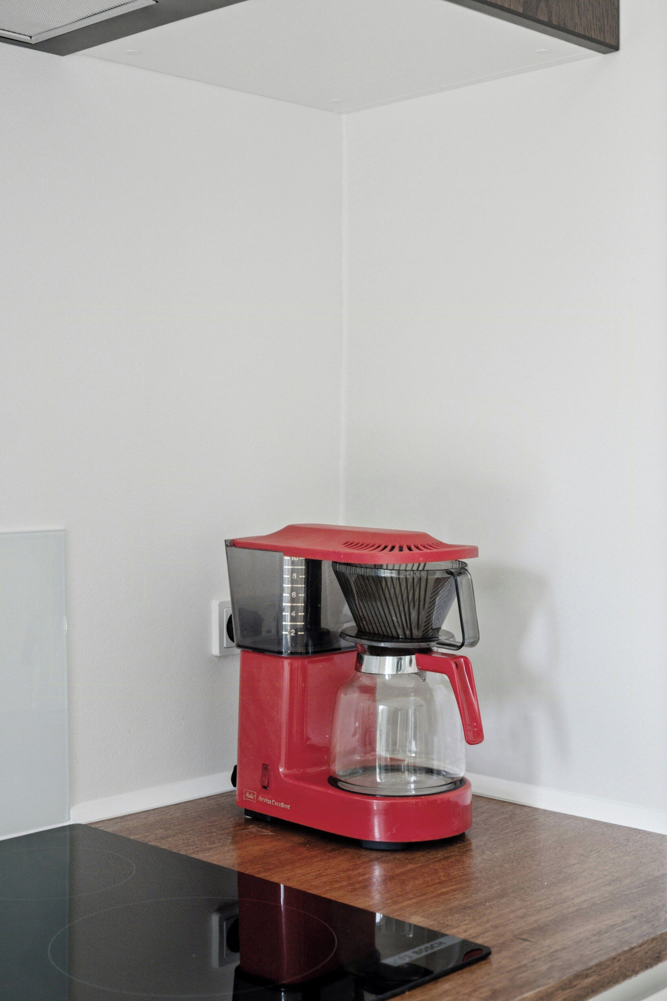 A red coffee maker on a wooden table against a white wall.