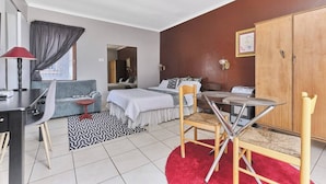 A hotel room with a bed, a desk, a chair, a small table, a wardrobe, and a window with curtains. - The Cozy 1 Bedroom Apartment (Johannesburg)
