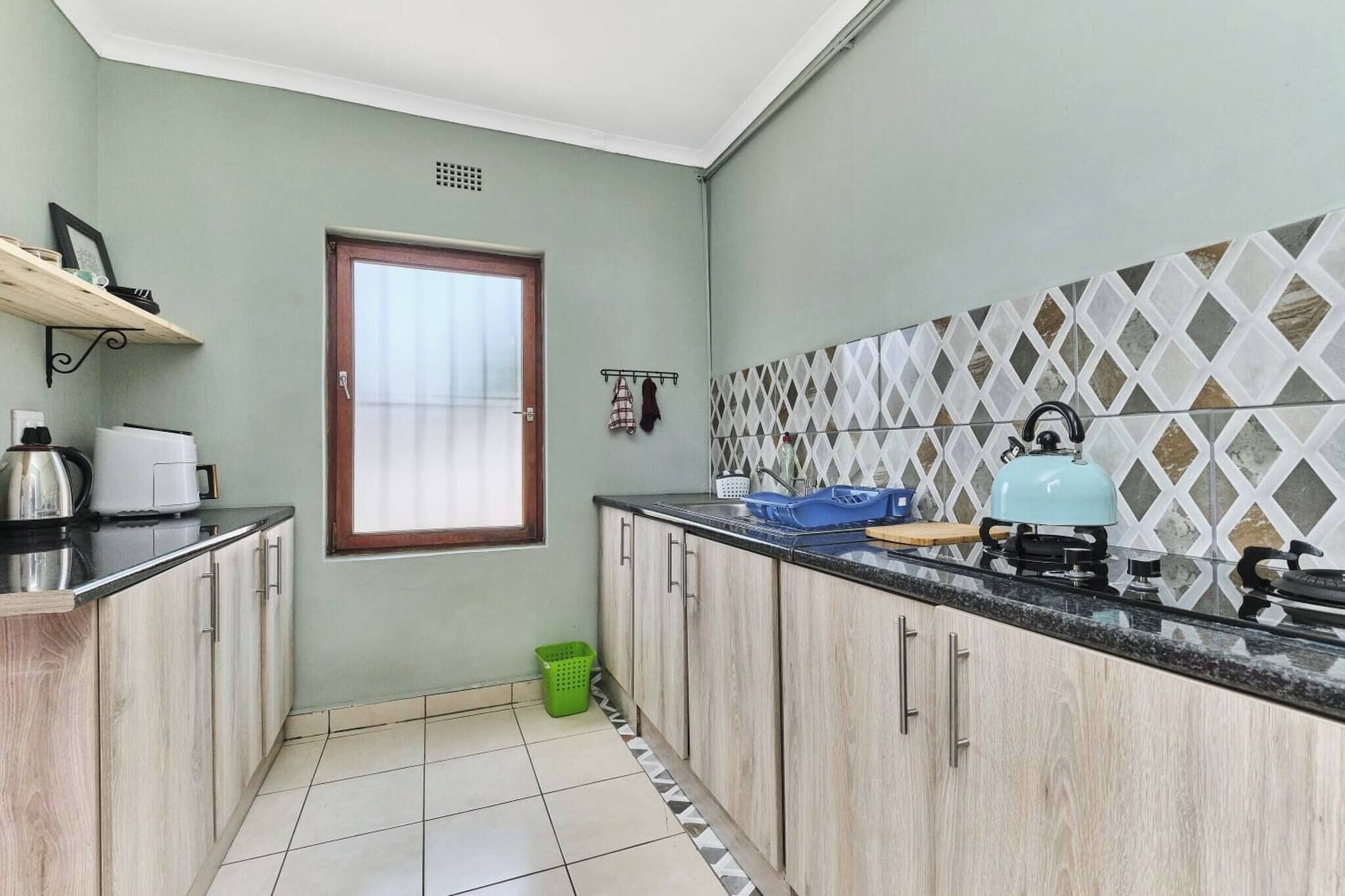 A kitchen with light green walls, a window, a tiled backsplash, and a green basket on the floor.