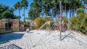 Ein sandiger Strand mit einer Holz-Spielkonstruktion, einer Seilbrücke und einem kleinen Holzgebäude.