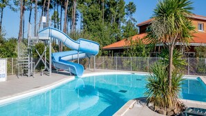 Ein Swimmingpool mit einer blauen Rutsche, umgeben von einem Zaun, Bäumen und einem Gebäude mit rotem Dach.