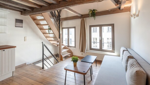 A loft-style room with exposed wooden beams, a staircase, a sofa, a small table, and a window with curtains. - Spacious 11-person Stay in Antwerp (Antwerp)