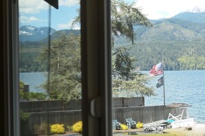 A view through a window of a lake with mountains in the background, a wooden dock, and a flag. - World Cup? Beachfront suite, 0.5 hr + ferry ride to Seattle. (Seabeck)