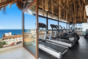 Ein Fitnessraum mit Laufbändern und Trainingsgeräten, große Fenster mit Blick auf das Meer und Gebäude.