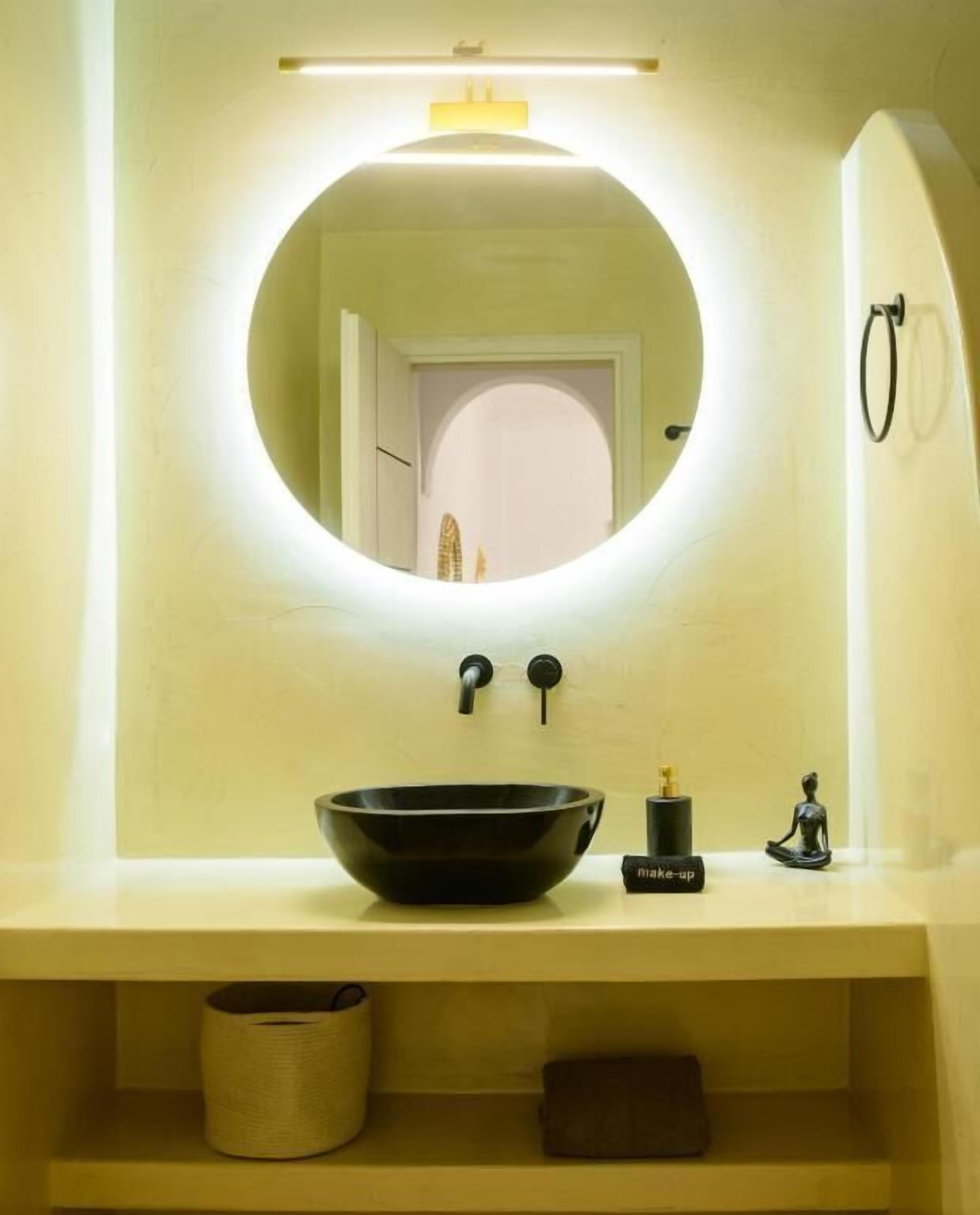 Un baño con un espejo redondo, un lavabo de cerámica negra, una pequeña estatua y una botella con la etiqueta 'maquillaje'.
