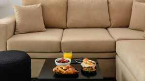 Ein beiges Sofa mit vier Kissen, ein schwarzer Hocker und ein Tisch mit Frühstücksartikeln, darunter Croissants, eine Fruchtschale, ein Glas Orangensaft sowie ein Teller mit Eiern und Gemüse.