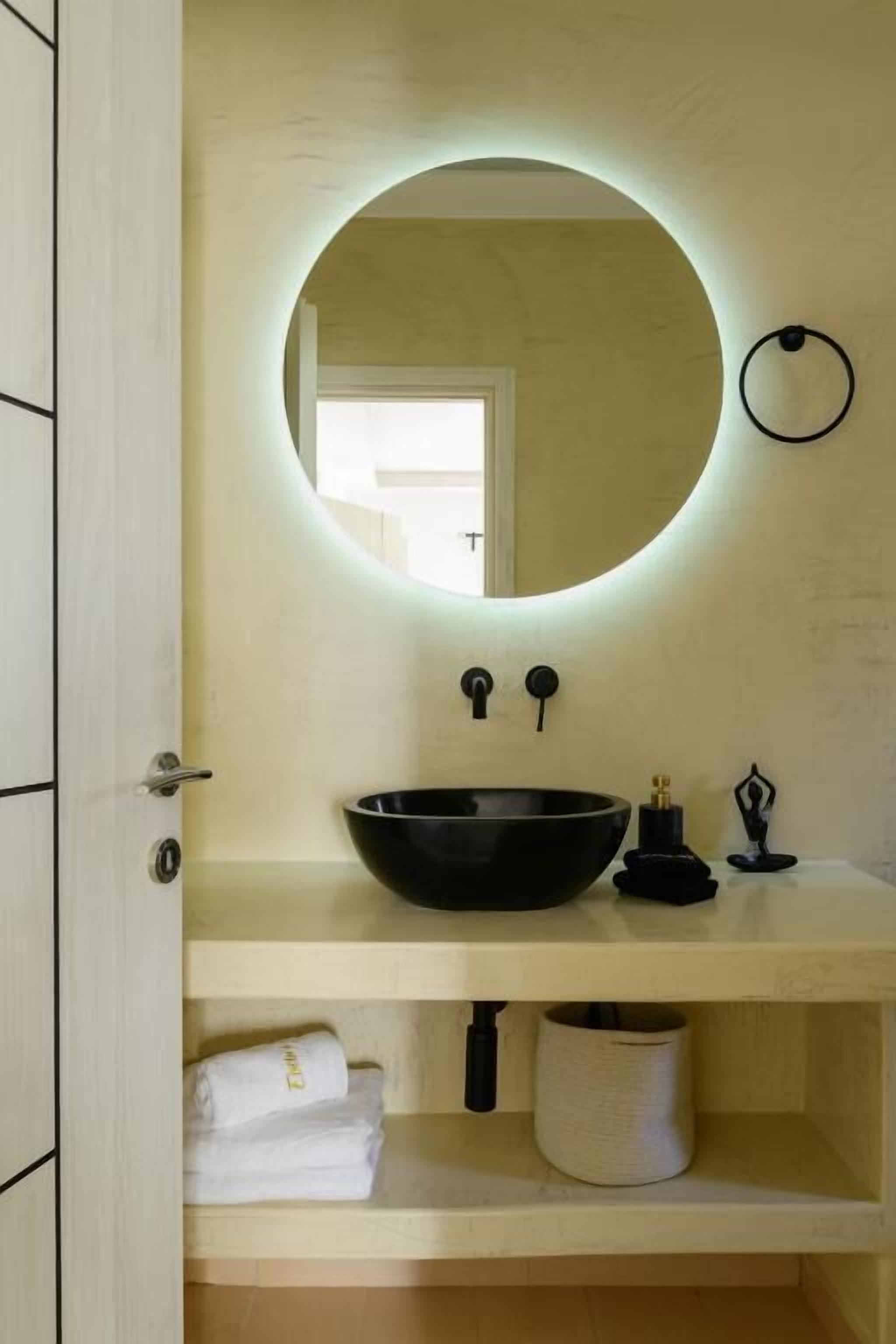 Un baño con un espejo redondo, un lavabo negro, una toalla y un estante pequeño con un recipiente.