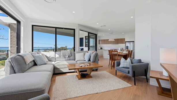 A modern living room with a large sofa, a coffee table, and a dining area with a table and chairs. - Panoramic Point by Experience Jervis Bay (Vincentia)