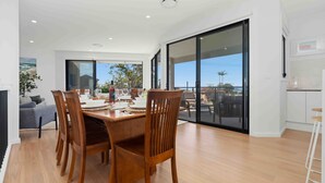 A dining area with a wooden table, chairs, and a view through large glass doors. - Panoramic Point by Experience Jervis Bay (Vincentia)