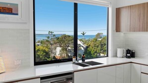 A kitchen with a large window, white cabinets, and a view of the ocean and trees. - Panoramic Point by Experience Jervis Bay (Vincentia)
