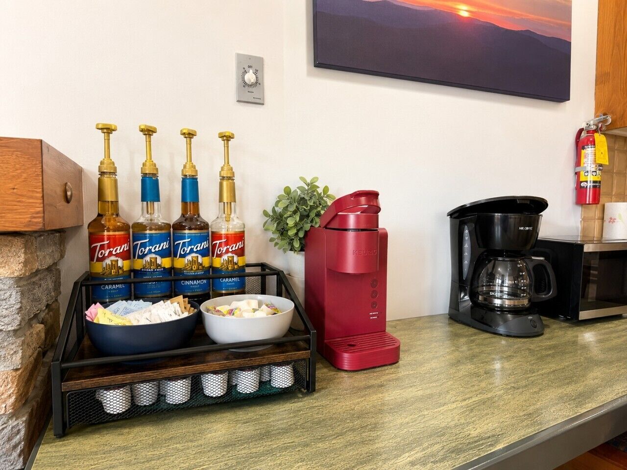 Una encimera de cocina con botellas de sirope Torani, una cafetera Keurig roja, un cuenco de aperitivos y una máquina de café.