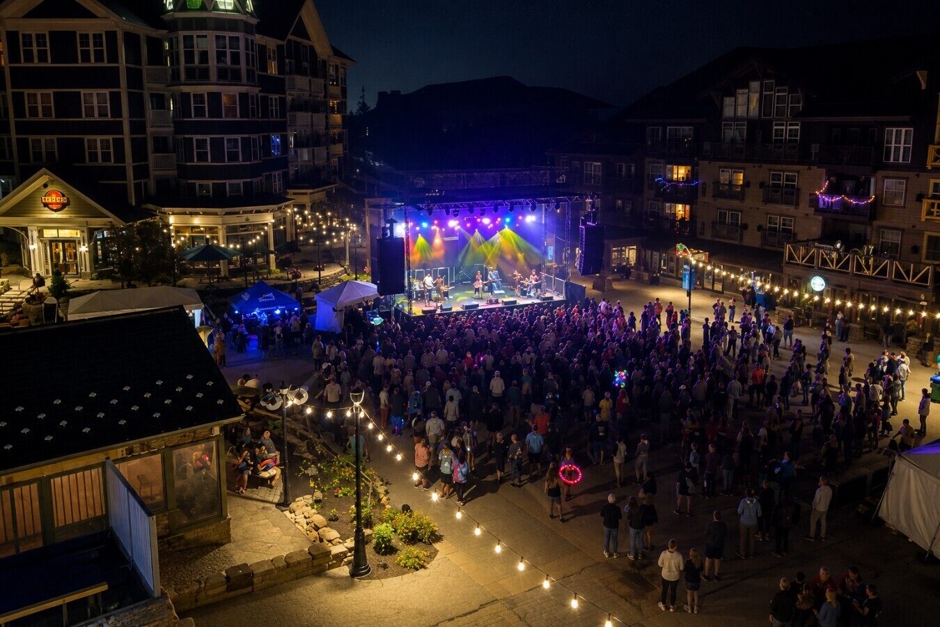 A large outdoor gathering with a stage and audience, surrounded by buildings with balconies and string lights.