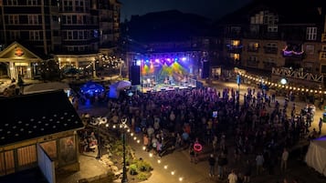 A large outdoor gathering with a stage and audience, surrounded by buildings with balconies and string lights.