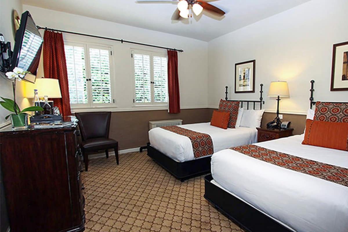 A hotel room with two beds, a desk, a chair, a desk lamp, a floor lamp, and a ceiling fan.