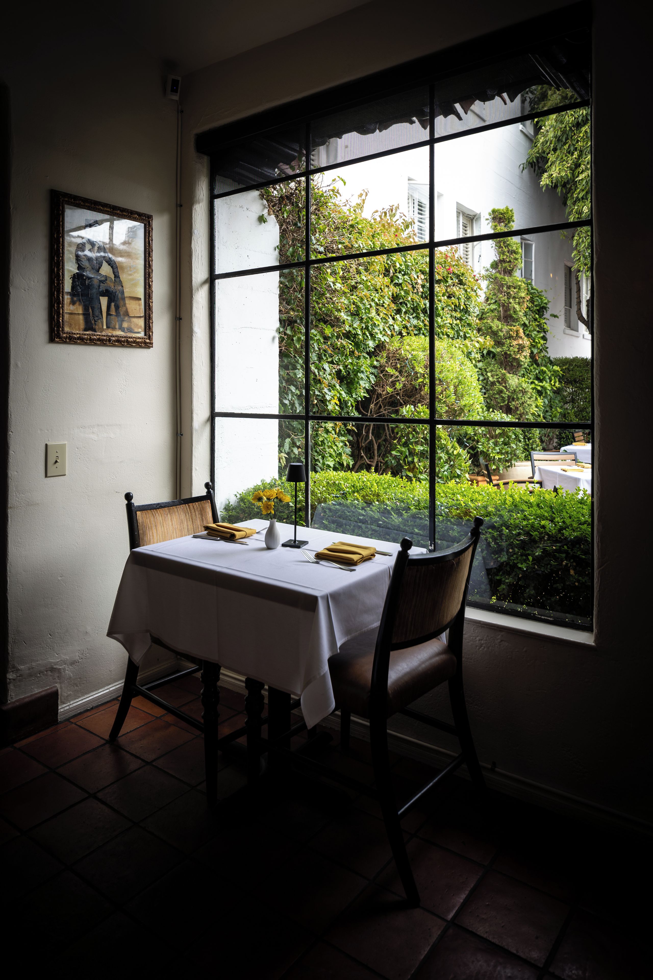 A dining area with a table set for two, a chair, a vase with flowers, and a painting on the wall.