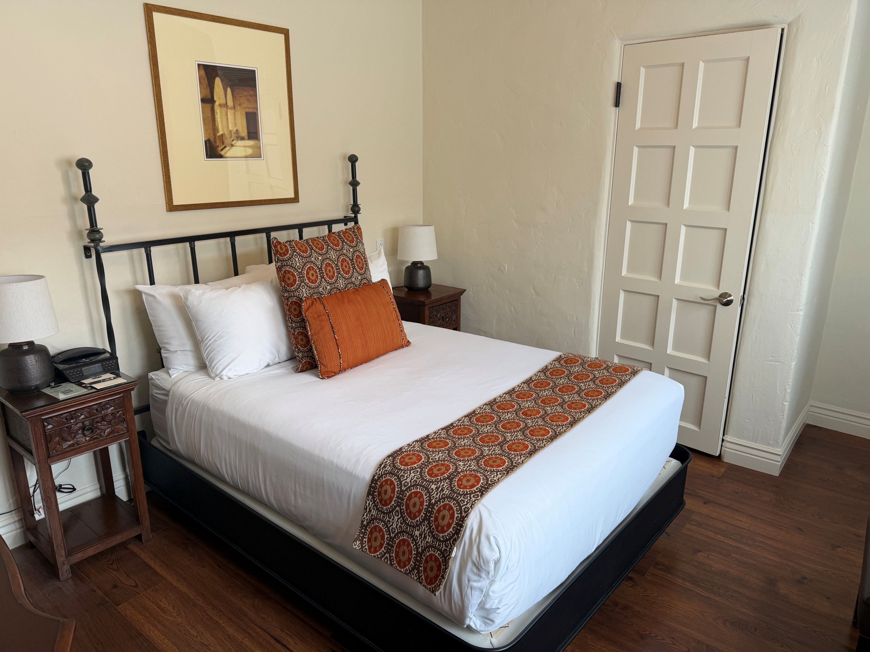 A neatly made bed with a patterned bedspread, a framed picture on the wall, a bedside lamp, and a door with a simple design.