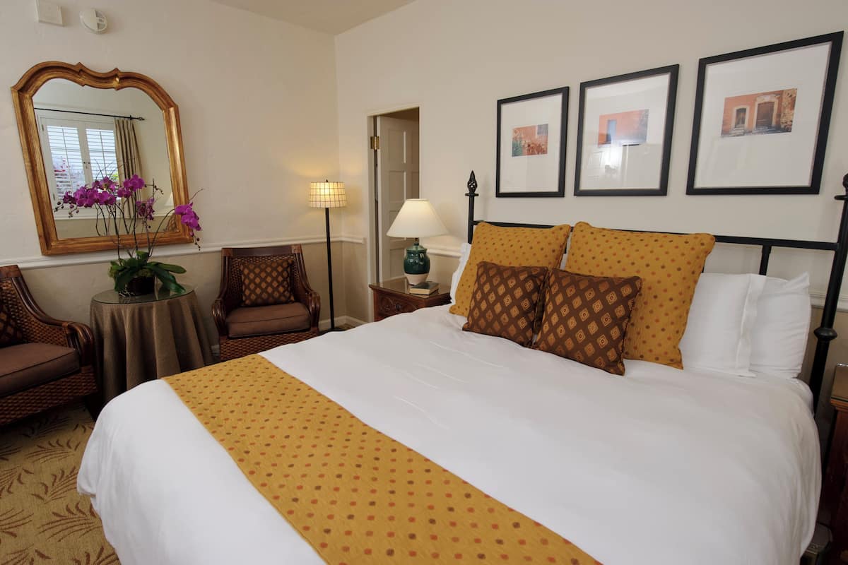 A hotel room with a large bed, a mirror, a small table with a lamp, a chair, and framed pictures on the wall.
