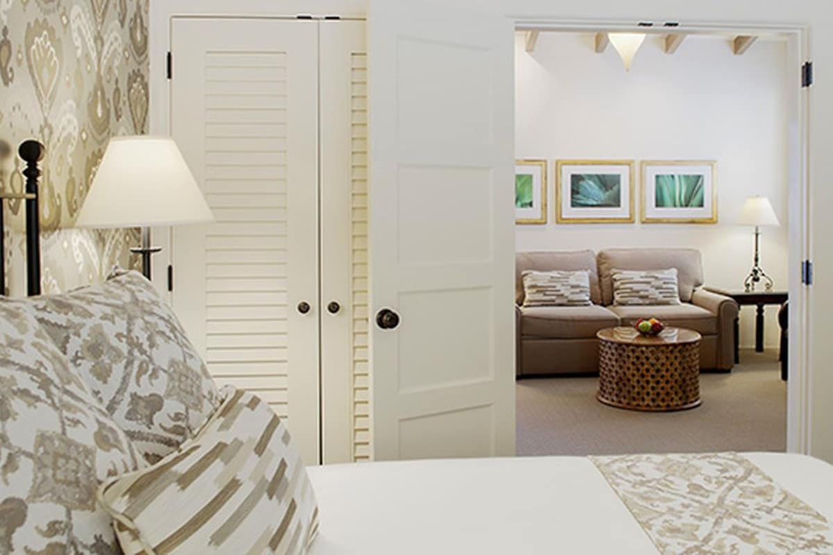 A hotel room with a bed, a sofa, a coffee table, and a door leading to a living area with a couch and framed pictures.