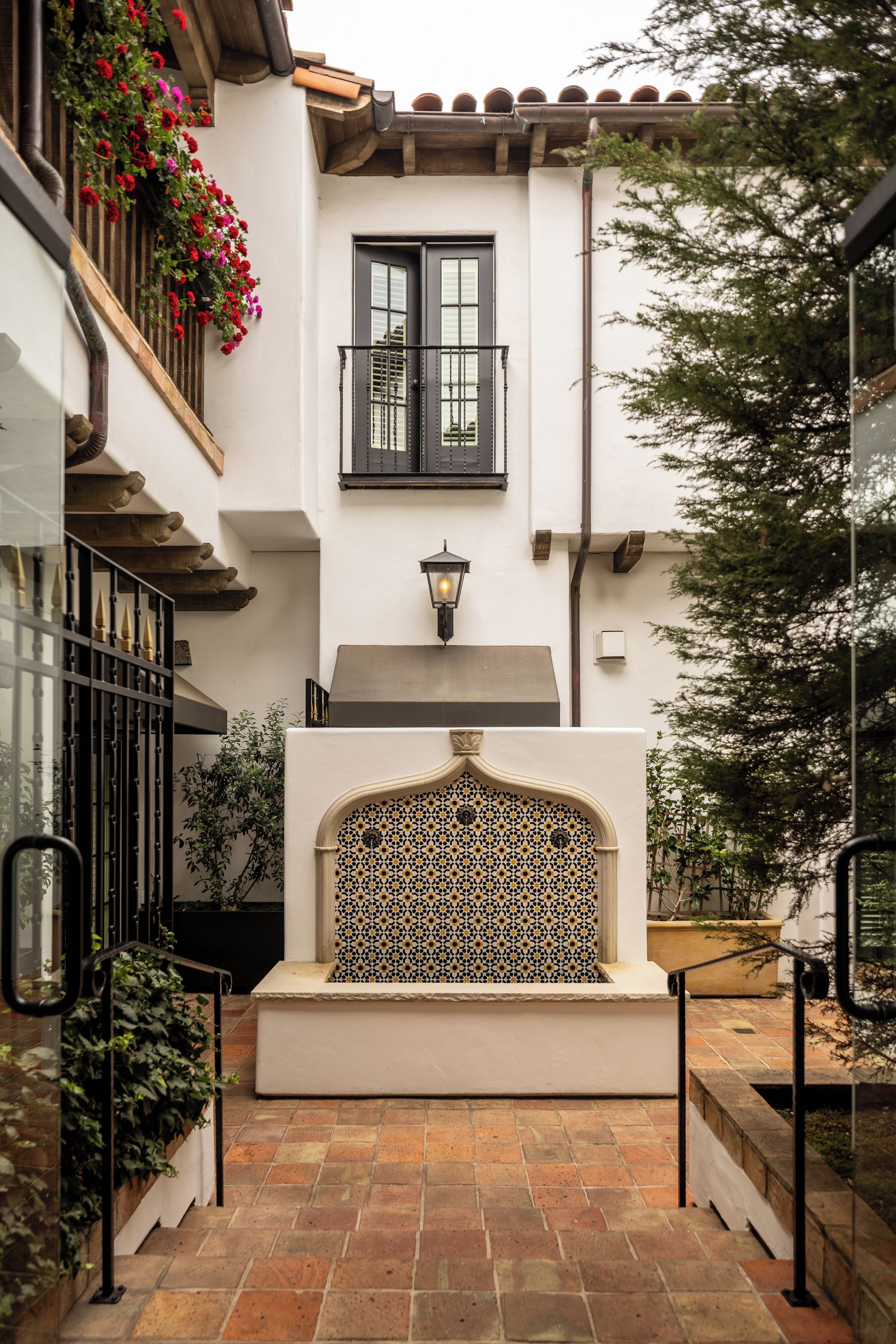Um pátio com um muro de azulejos, uma varanda florida e uma escadaria que leva a uma porta.