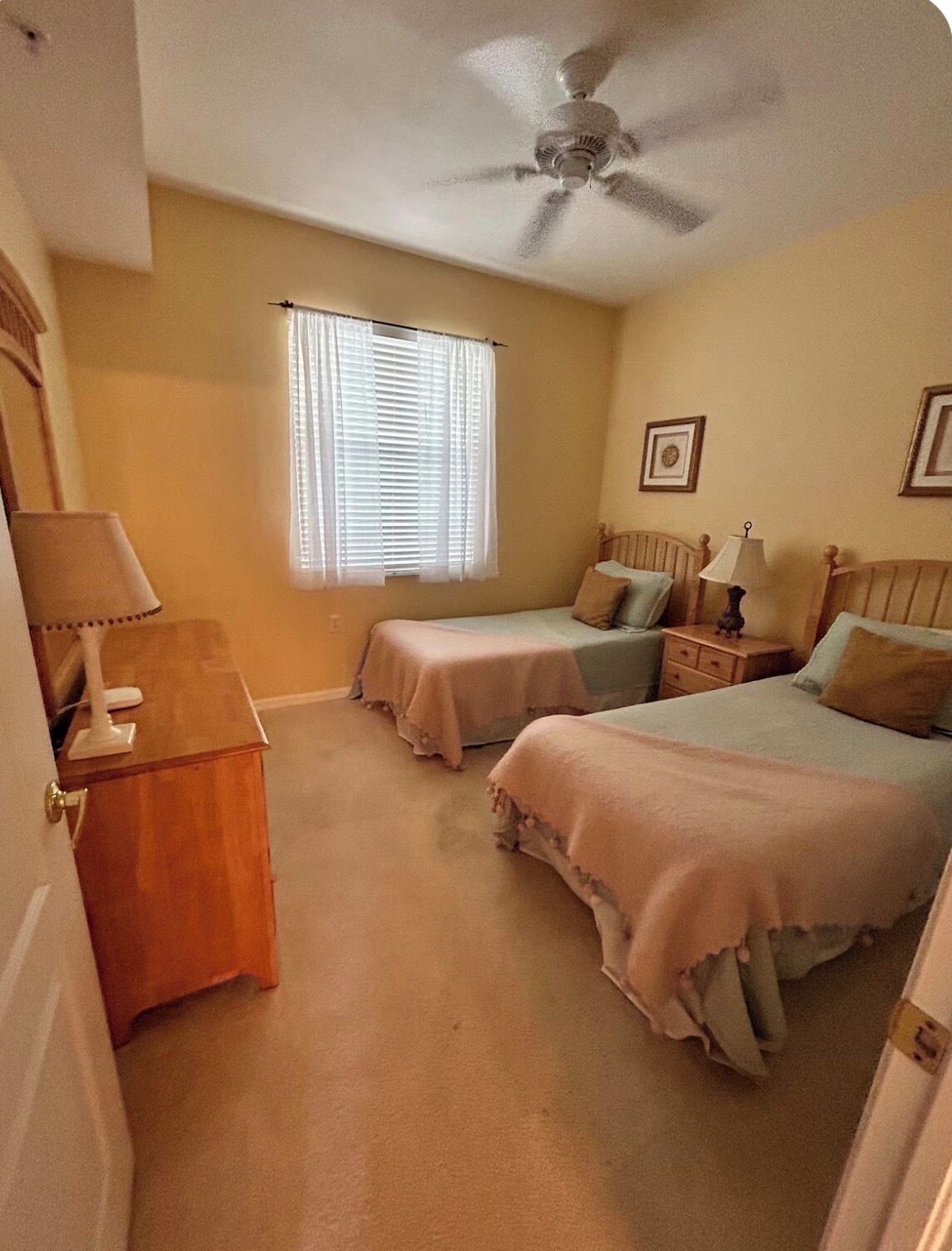 A bedroom with two single beds, a ceiling fan, a window with blinds, a bedside table with a lamp, and a wooden dresser.