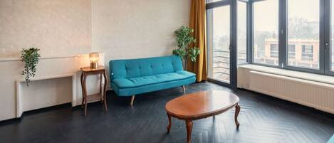 A room with a blue sofa, a wooden table, a floor lamp, and a window with a view of buildings.