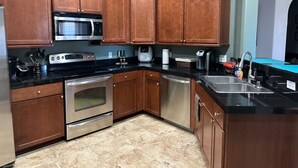 Una cocina con armarios de madera, electrodomésticos de acero inoxidable, una encimera negra y suelo de baldosas.