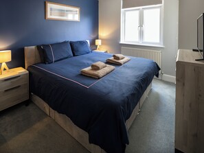 A neatly made bed with a dark blue bedspread and two towels, a bedside table with a lamp, a framed picture on the wall, and a window with blinds. - Woolacombe Apartment with Panoramic Sea Views (Woolacombe)