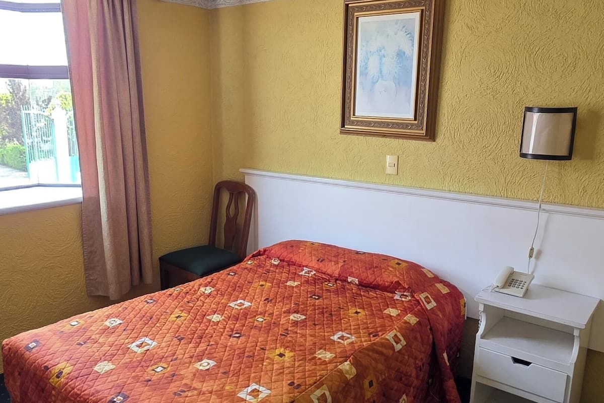 Une chambre d’hôtel avec un lit, une table de chevet, un téléphone, un cadre photo et une fenêtre avec des rideaux.
